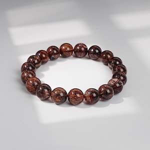 May include: A stretch bracelet composed of round, polished beads made of dark reddish-brown rutilated quartz. Each bead features distinct, needle-like inclusions that create a shimmering, fibrous texture within the translucent, deep-toned stone material.