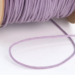 May include: A spool of lavender-colored waxed cotton cord. The cord is wound tightly around the spool and a length of cord is hanging off the spool.