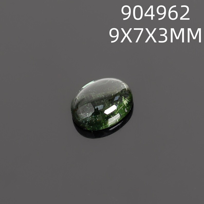 May include: A polished, oval-shaped gemstone with a dark green hue and internal inclusions. The gem measures 9x7x3mm and is set against a dark gray background. The text "904962 9X7X3MM" is displayed above the gem.