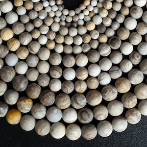 May include: A collection of small, round, stone beads in various shades of gray, brown, and cream. The beads have a marbled or striped appearance, with some displaying a more solid color. The arrangement is dense, with the beads closely packed together.