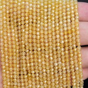 Puede incluir: Primer plano de hebras de peque&ntilde;as cuentas facetadas de color amarillo dorado. Las cuentas est&aacute;n ensartadas, creando un efecto brillante. Las cuentas se sostienen en una mano, mostrando su color y textura. Ideal para la fabricaci&oacute;n de joyas o manualidades.