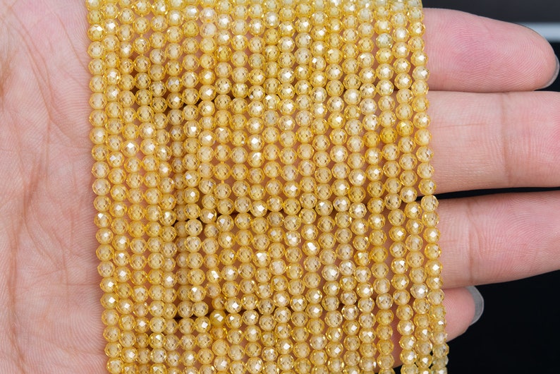 Puede incluir: Primer plano de hebras de peque&ntilde;as cuentas facetadas de color amarillo dorado. Las cuentas est&aacute;n ensartadas, creando un efecto brillante. Las cuentas se sostienen en una mano, mostrando su color y textura. Ideal para la fabricaci&oacute;n de joyas o manualidades.