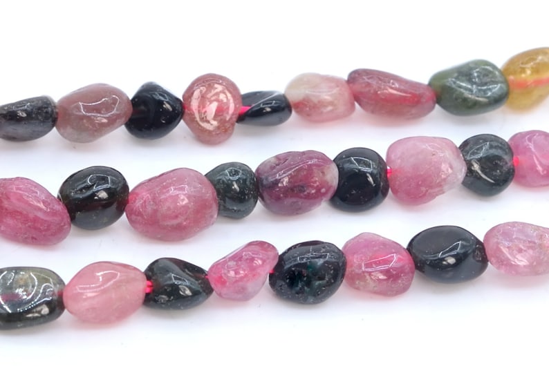 May include: A strand of multi-colored gemstone beads. The beads are a mix of pink, black, and green, and are tumbled into a smooth, oval shape.