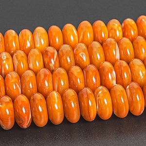 Op de afbeelding: Rijen gepolijste, ovale barnsteen kralen. De kralen hebben een warme oranje tint met variaties in kleur en interne patronen. De kralen zijn dicht op elkaar geplaatst, waardoor een textuur ontstaat. De achtergrond is donker.