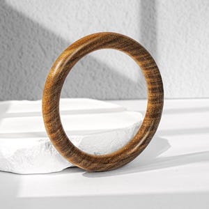 May include: A circular bangle bracelet crafted from polished brown wood, featuring a distinct natural grain pattern with swirling dark and light brown tones throughout the smooth, rounded surface of the solid wooden ring structure.