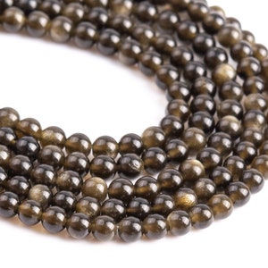 Puede incluir: Primer plano de hebras de cuentas de piedras preciosas con brillo dorado. Las cuentas son redondas, con un color base marr&oacute;n oscuro y motas doradas brillantes. Las cuentas est&aacute;n ensartadas, para la creaci&oacute;n de joyas. Fondo blanco.