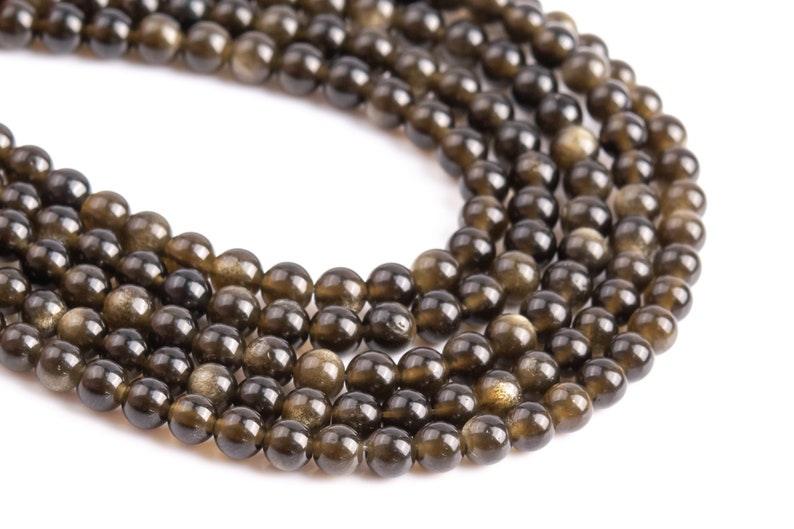 Puede incluir: Primer plano de hebras de cuentas de piedras preciosas con brillo dorado. Las cuentas son redondas, con un color base marr&oacute;n oscuro y motas doradas brillantes. Las cuentas est&aacute;n ensartadas, para la creaci&oacute;n de joyas. Fondo blanco.