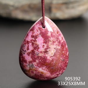May include: A teardrop-shaped pendant with a pink and maroon marbled pattern. The gemstone is suspended by a maroon cord. The pendant measures 33mm x 25mm x 8mm.