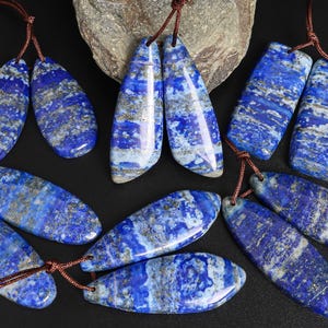 May include: A collection of polished lapis lazuli pendants in various shapes, including teardrop and rectangular. The deep blue stones feature streaks of white and gold, each strung on a brown cord. The pendants are displayed on a black surface.