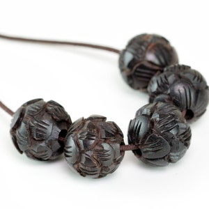 Puede incluir: Un collar de madera marr&oacute;n oscuro con cuentas talladas. Las cuentas son redondas y tienen un patr&oacute;n floral. El collar est&aacute; encordado en un cord&oacute;n marr&oacute;n.