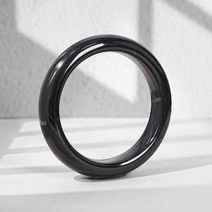 May include: A smooth, circular bangle bracelet with a polished, glossy black finish. The bangle features a rounded, thick profile and a solid, uniform appearance, reflecting light across its curved surface to highlight its dark, deep color.