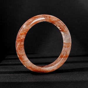 May include: A circular bangle bracelet crafted from polished red rutilated quartz. The stone features a translucent, clear base with prominent, deep red needle-like inclusions that create a complex, natural pattern throughout the entire band.