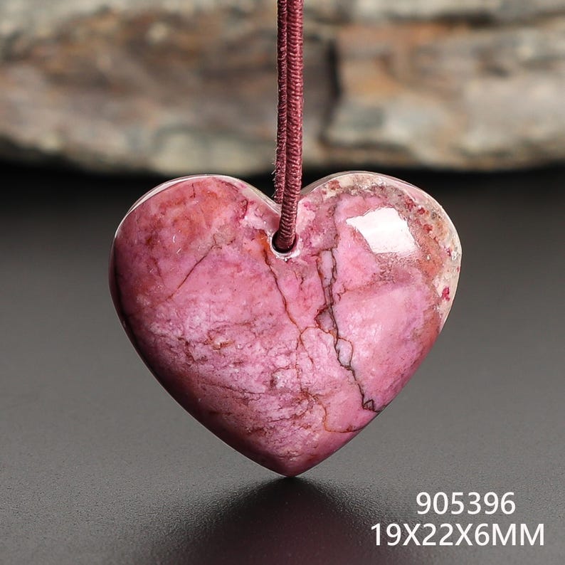 May include: A heart-shaped pendant made of pink stone, suspended by a maroon cord. The stone has a marbled appearance with darker veins. The pendant measures 19x22x6mm. The text "905396" is visible.