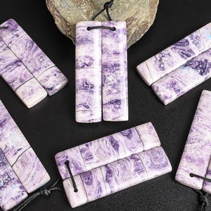 May include: A collection of rectangular pendants with a marbled purple and white pattern. Each pendant is connected to a black cord. The pendants are arranged on a black surface, with a rock in the background.