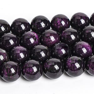 May include: A strand of dark purple gemstone beads with a shiny, iridescent finish. The beads are round and smooth, and they have a subtle sparkle.