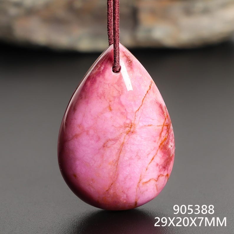 May include: A teardrop-shaped pendant with a pink and red marbled pattern, suspended by a maroon cord. The pendant measures 29mm x 20mm x 7mm. The surface is polished and smooth, with natural veining.