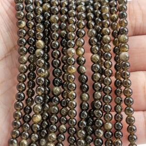 Puede incluir: Hebras de cuentas de piedras preciosas de obsidiana dorada. Las cuentas son peque&ntilde;as, redondas y tienen un aspecto oscuro y reflectante con motas doradas. Las cuentas est&aacute;n ensartadas, creando un efecto brillante. El fondo es un tono de piel claro.