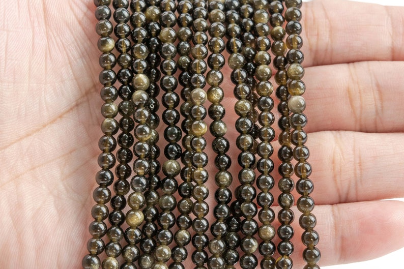 Puede incluir: Hebras de cuentas de piedras preciosas de obsidiana dorada. Las cuentas son peque&ntilde;as, redondas y tienen un aspecto oscuro y reflectante con motas doradas. Las cuentas est&aacute;n ensartadas, creando un efecto brillante. El fondo es un tono de piel claro.