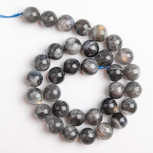 May include: Strand of round, polished gemstone beads in shades of gray, black, and silver. The beads are strung on a blue cord, creating a necklace or bracelet component. The beads are approximately 10mm in diameter.