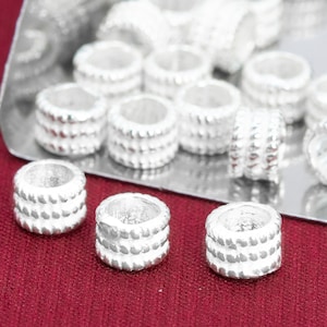 May include: A close-up of a group of silver-toned metal beads with a textured design. The beads are arranged on a silver surface and a red surface.