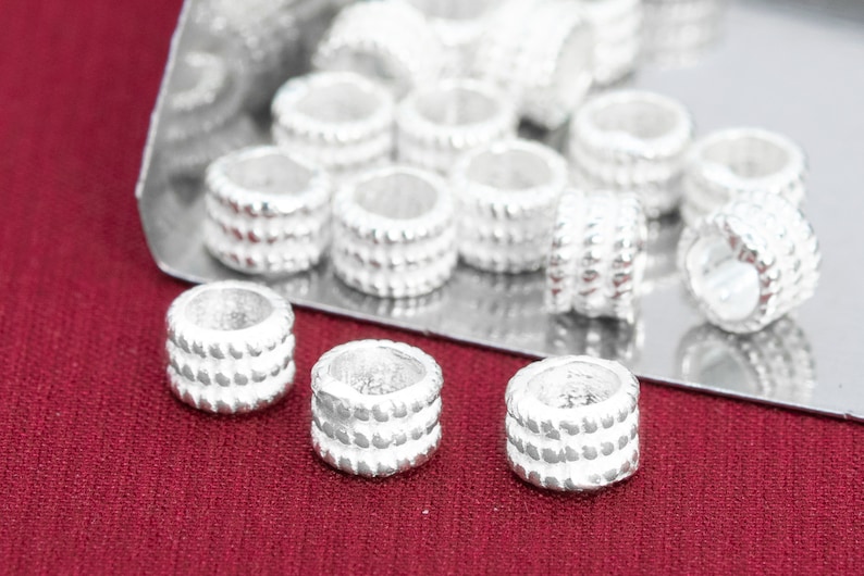 May include: A close-up of a group of silver-toned metal beads with a textured design. The beads are arranged on a silver surface and a red surface.