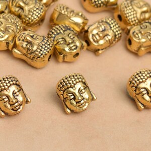 May include: A group of gold-colored metal beads with a Buddha face design. The beads are scattered on a light brown surface.