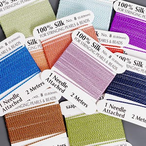 Puede incluir: Una colección de carretes de hilo de seda en varios colores, incluyendo azul, naranja y morado. Cada carrete tiene una etiqueta que dice "100% Seda" y "Para ensartar perlas y abalorios". Las etiquetas también indican que se adjunta una aguja y que el hilo mide 2 metros.