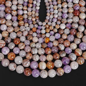 May include: A collection of gemstone beads in various colors, including purple, brown, and white. The beads are round and have a polished surface, with natural patterns and textures. The beads are arranged closely together, creating a dense, textured surface.
