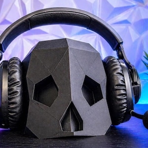 Op de afbeelding: Zwarte gamingheadset op een zwarte schedelvormige hoofdtelefoonstandaard. De headset heeft grote oorschelpen en een boommicrofoon. De standaard heeft een geometrisch ontwerp. De achtergrond is een getextureerde muur met blauwe en witte verlichting.