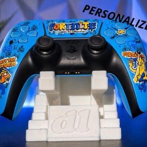 Op de afbeelding: Een blauwzwarte Fortnite-thema videogamecontroller met een witte standaard. De controller heeft verschillende cartoonafbeeldingen en het woord "Fortnite" in het geel. De standaard heeft de letters "d1" en het woord "GEPERSONALISEERD" in het zwart.
