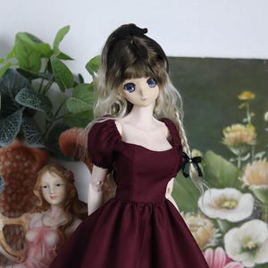 May include: A burgundy doll dress with puffed sleeves and a square neckline. The dress has a fitted bodice and a full skirt. The doll is wearing a black bow in her hair.