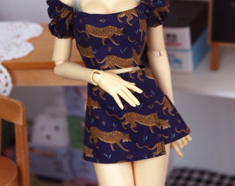 May include: A doll wearing a navy blue two-piece outfit with a gold leopard print. The doll has pale skin, blue-gray wavy hair, and is wearing a silver necklace. The outfit includes a crop top and a mini skirt.