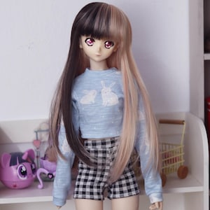 May include: A doll with long black and pink hair wearing a light blue crop top with a white bunny print and black and white checkered shorts. The doll is standing in front of a white shelf with a pink toy on the shelf.