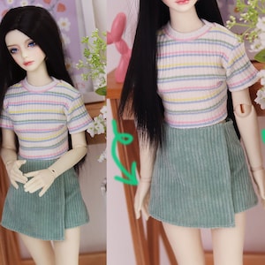 May include: A doll wearing a short-sleeved, ribbed crop top with horizontal stripes in pastel colors. The doll also wears a wrap-style, corduroy mini-skirt in a muted green color. The doll has long black hair and is posed indoors.