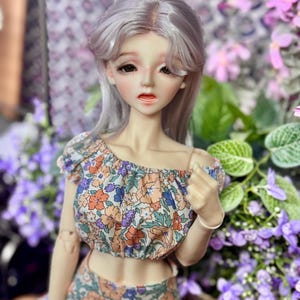 May include: A doll with gray hair and a floral crop top and skirt. The top is off-the-shoulder with a pattern of orange, purple, and blue flowers. The skirt has a matching floral pattern. The doll is surrounded by purple flowers.