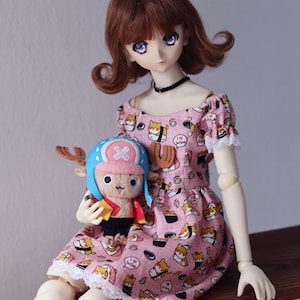 May include: A doll with brown hair and purple eyes wearing a pink dress with a sushi pattern. The doll is holding a plush toy of a character wearing a blue hat. The dress has white lace trim.