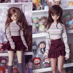 May include: Two dolls wearing striped long-sleeved shirts and burgundy skirts. The dolls have long brown hair and are standing in front of a shelf with other dolls.