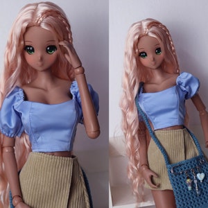 May include: A doll with long, wavy, pink hair wearing a light blue crop top with puffed sleeves and a beige corduroy skirt. The doll is holding a blue crocheted bag with charms.