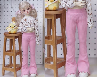 Peut inclure: Une poupée vêtue d'un haut blanc à motif d'ours marron et d'un pantalon évasé côtelé rose. La poupée porte également des baskets blanches. Un petit tabouret en bois et un poussin crocheté jaune sont en arrière-plan.