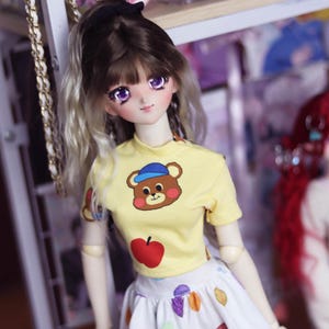 May include: A doll with long, blonde and brown hair, wearing a yellow shirt with a cartoon bear and a white skirt with colorful polka dots.