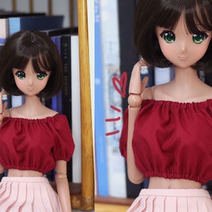 May include: A doll with green eyes and short brown hair, wearing a red off-the-shoulder top and a pink pleated skirt. The doll is posed in front of a bookshelf. The doll's arms are articulated.