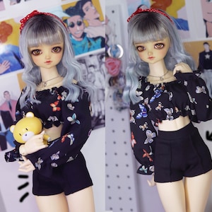 May include: A doll with long, silver hair wearing a black and white butterfly print off-the-shoulder top and black shorts. The doll is holding a yellow stuffed animal.