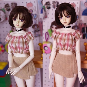 May include: A doll with a short, dark brown wig, wearing a pink and brown plaid off-the-shoulder crop top, a tan corduroy skirt, and a black choker. The doll has large, expressive eyes and is posed in front of a blurred background.