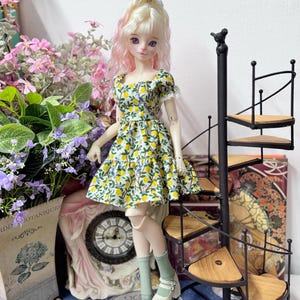May include: A doll wearing a yellow and green lemon print dress, light green socks, and white platform shoes. The doll has blonde hair with pink highlights and a blue bow. A black spiral staircase and floral arrangements are in the background.
