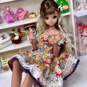 May include: A detailed doll with green eyes and blonde hair, wearing a dress with a teddy bear pattern. The dress has a blue lace trim. The doll is seated, with other dolls and collectibles visible in the background.