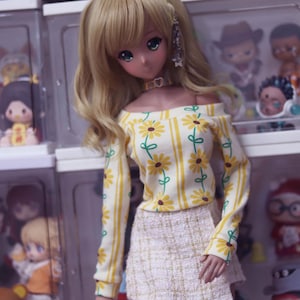 May include: A doll with blonde hair and green eyes, wearing a yellow off-the-shoulder top with a daisy pattern and a white tweed skirt. The doll is posed in front of a display case filled with other dolls.