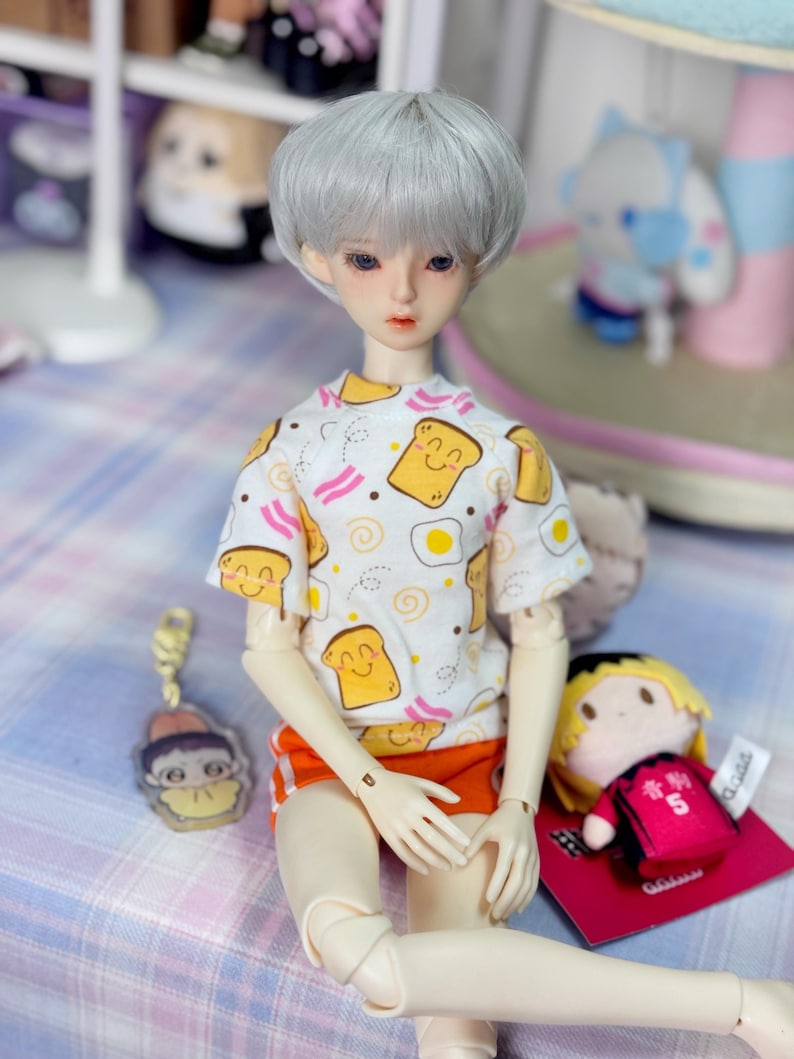 Puede incluir: Una mu&ntilde;eca con peluca plateada, que lleva una camiseta blanca con un estampado de desayuno y pantalones cortos naranjas. Un peque&ntilde;o llavero y un peluche est&aacute;n al lado de la mu&ntilde;eca. La mu&ntilde;eca est&aacute; sentada sobre una superficie a cuadros.