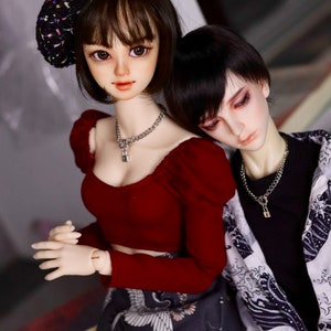 May include: Two dolls, one wearing a red top, a black and white skirt, and a black beret with colorful accents, and the other wearing a black shirt with a white and gray patterned shirt over it. Both dolls are wearing silver necklaces with lock pendants.