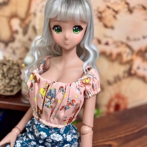 May include: A doll with silver hair and green eyes wearing a pink top with a cartoon print and a blue floral skirt. The doll is posed on a wooden surface, with a blurred background.