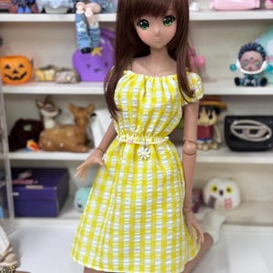 May include: A doll with brown hair and green eyes wearing a yellow and white gingham dress with a floral belt. The doll is kneeling. Shelves in the background display various figurines and toys.